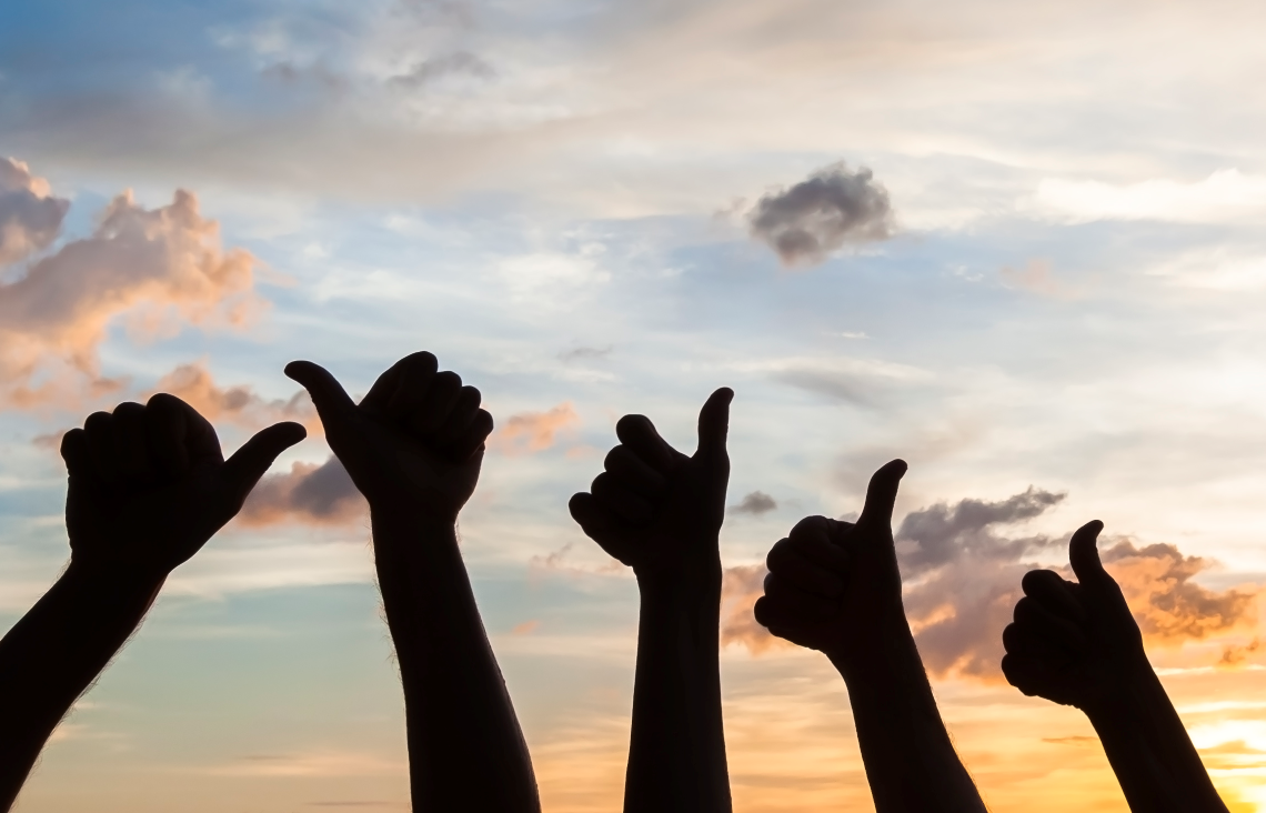 Hender som viser "tommmel opp"-tegnet mot en himmel i solnedgang